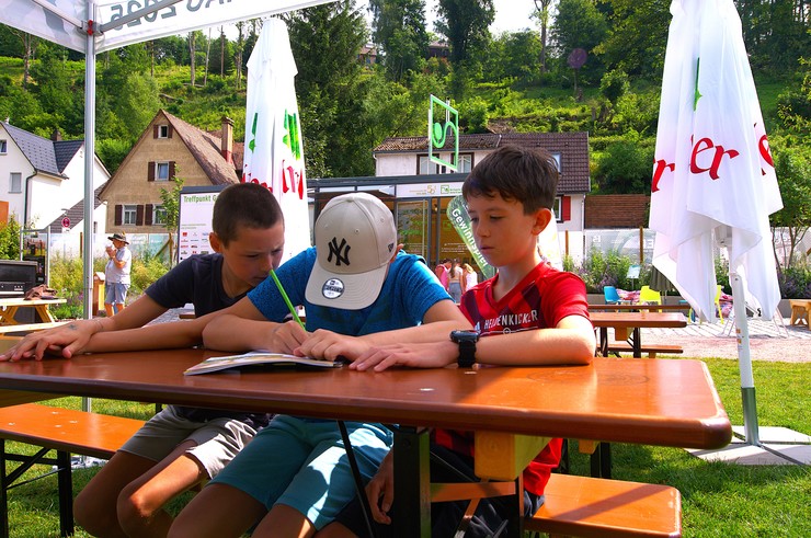 Bild: Drei Kinder sitzen an einer Biertischgarnitur im Freien und sind dabei Aufgaben zu lösen.